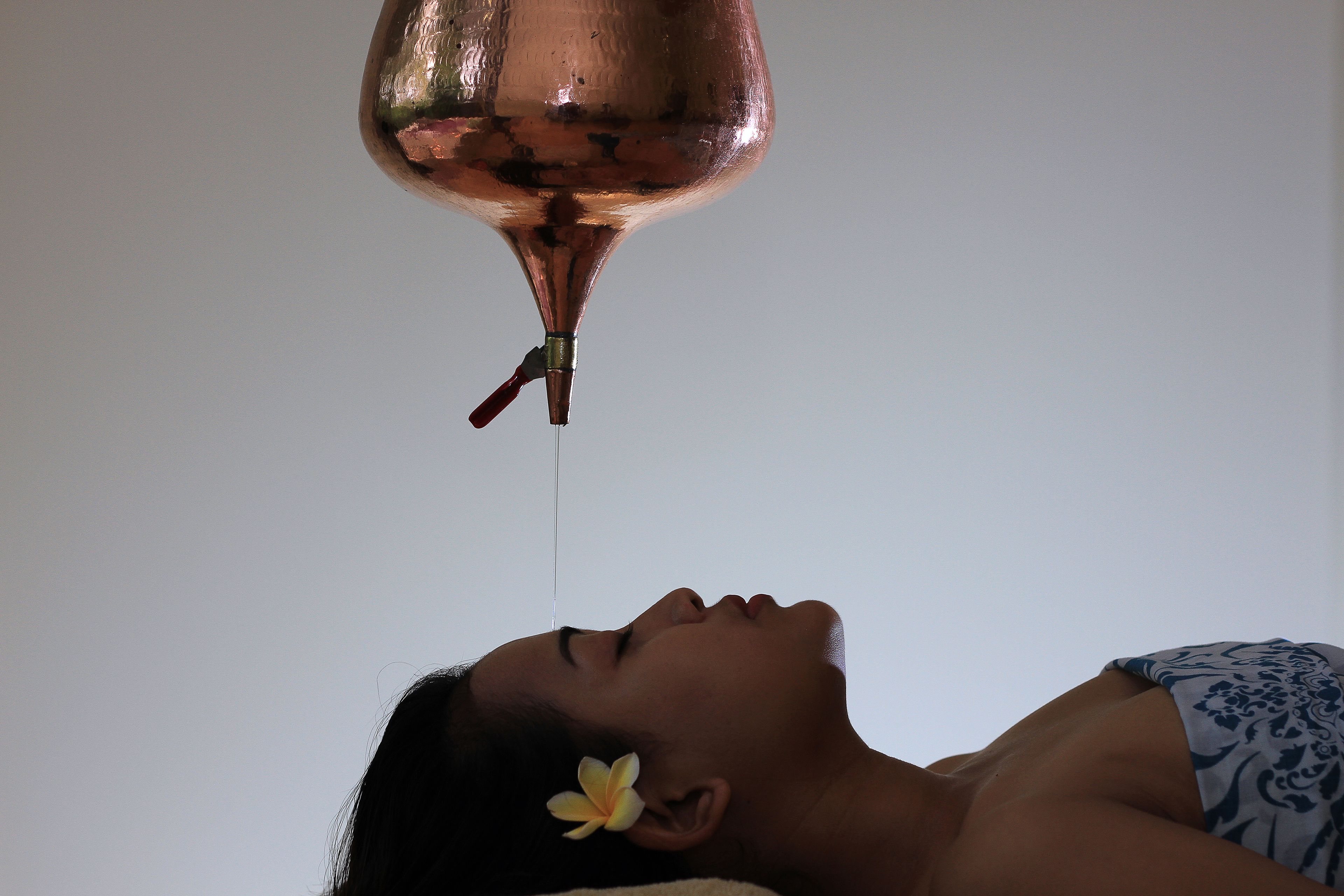 A person lying down with a flower in their hair, a copper vessel above them, and a white wall in the background.