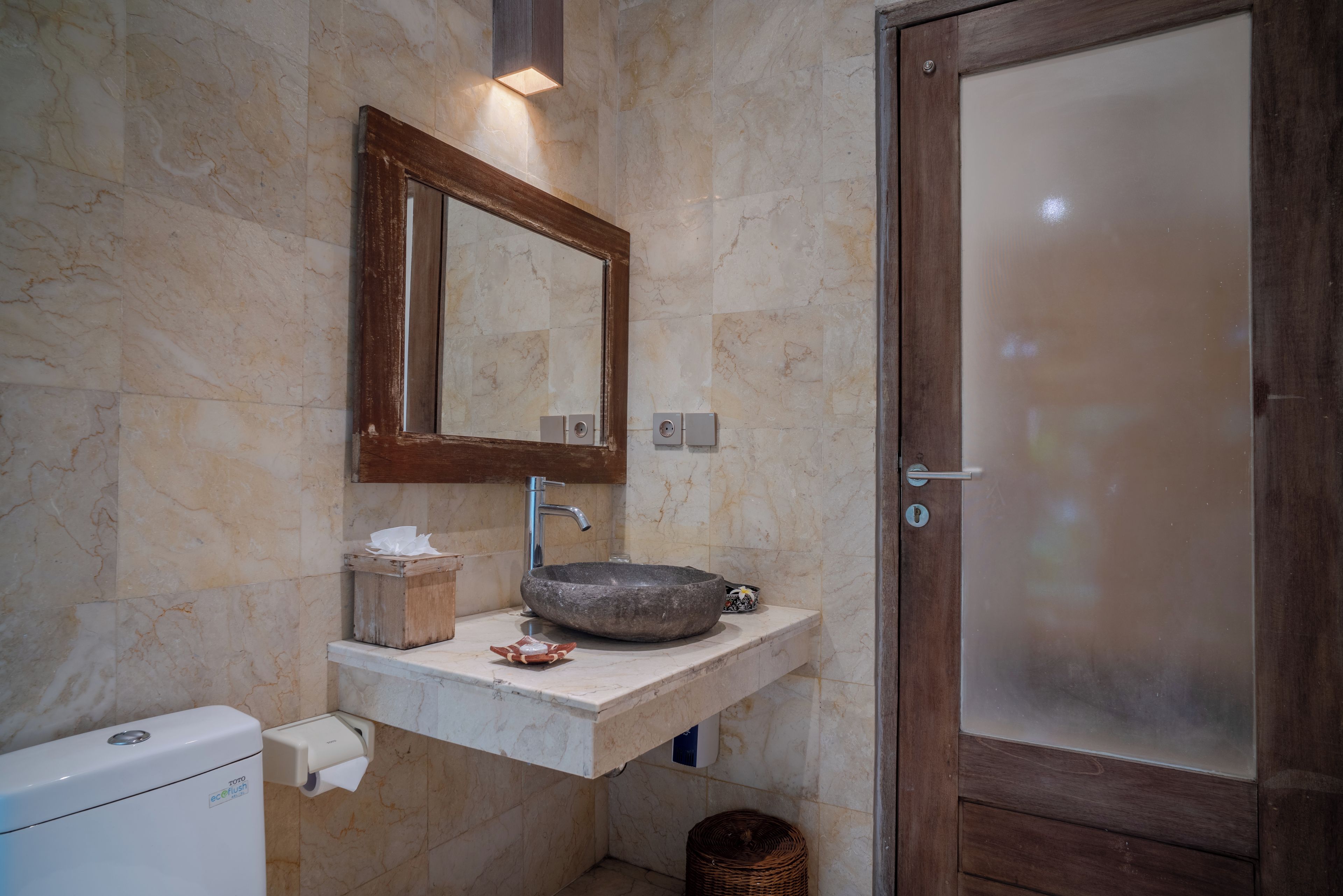 Une salle de bain avec un mur en marbre, un miroir encadré en bois, un lavabo sur colonne et des toilettes.