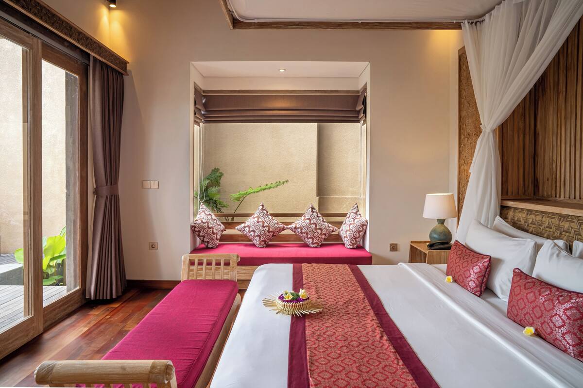 Une chambre d’hôtel avec un grand lit, un banc en bois, une petite table avec une lampe et une vue sur un balcon agrémenté de plantes.