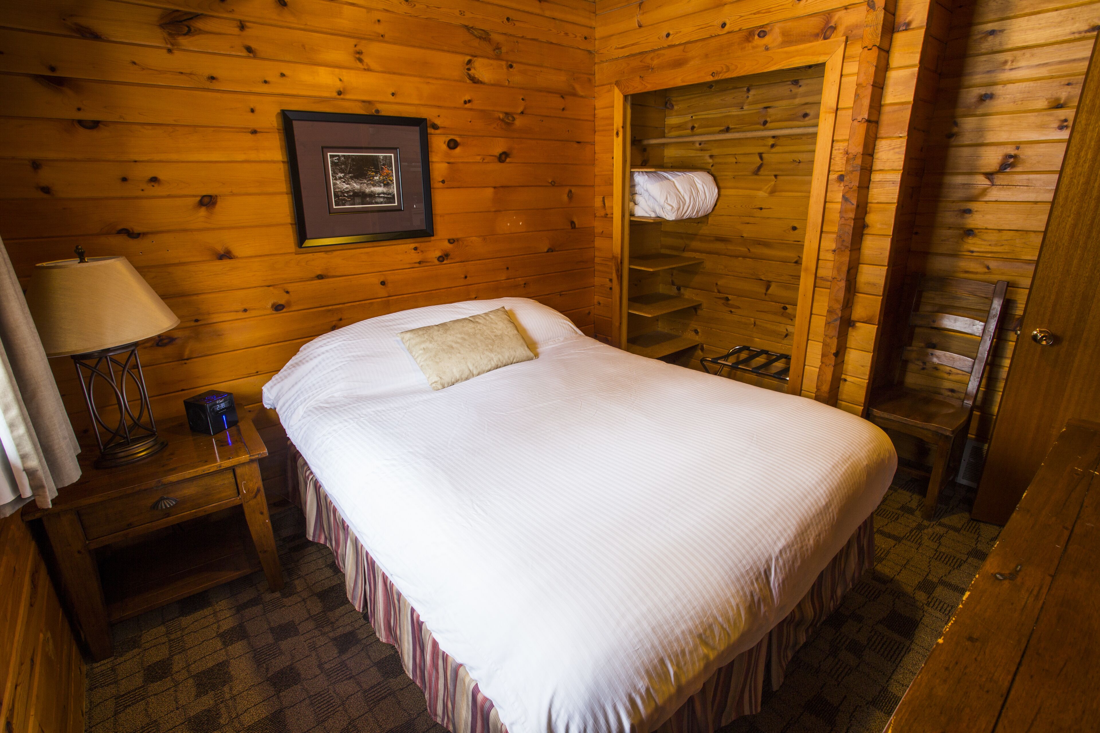 Une chambre à coucher dans une cabane en bois, avec un lit, une table de chevet dotée d’une lampe et un tableau encadré accroché au mur.