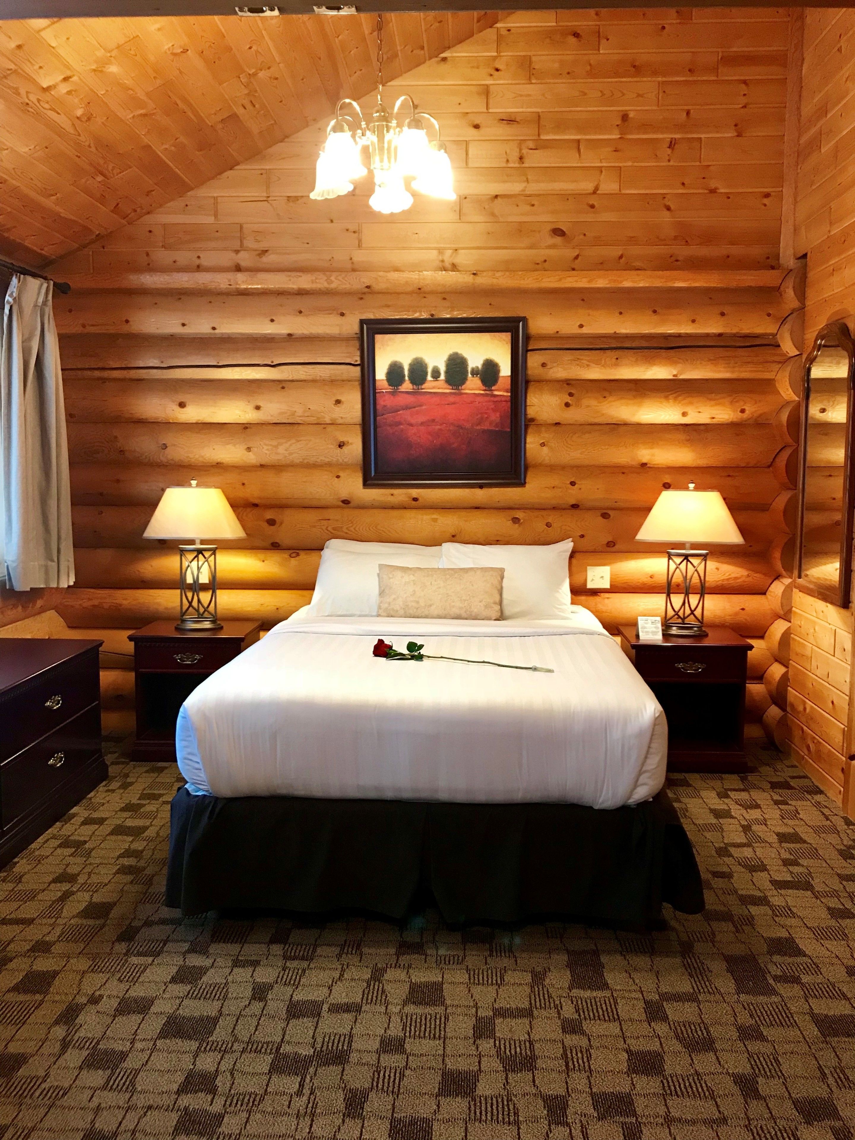 Une chambre confortable dans un chalet en bois, avec un grand lit, deux lampes de chevet, un tableau représentant un paysage encadré et un lustre.