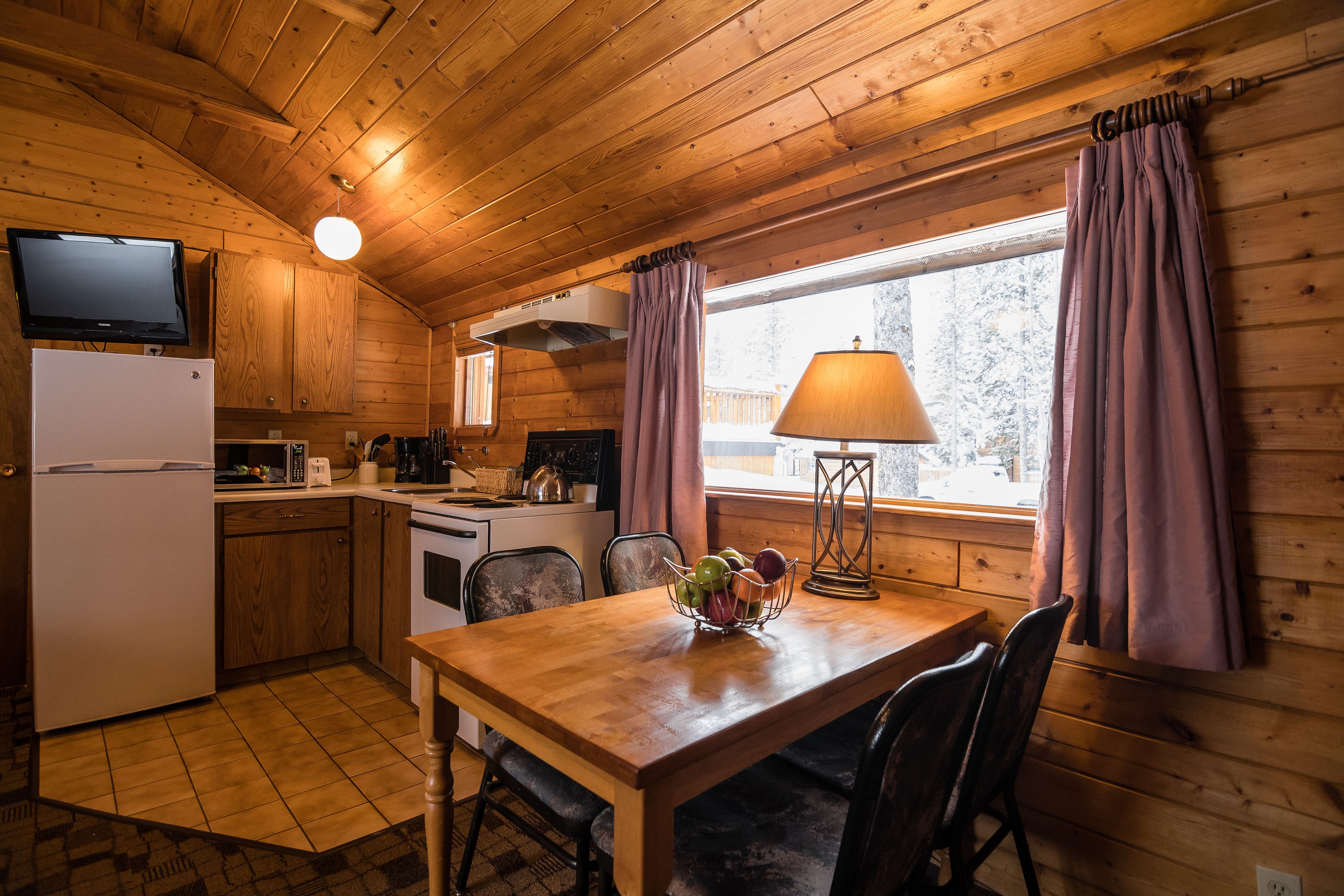 A wooden cabin interior with a kitchen, dining area, and a window with curtains.