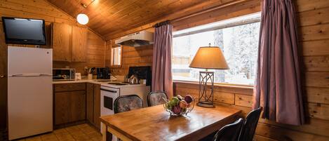 A wooden cabin interior with a kitchen, dining area, and a window with curtains.