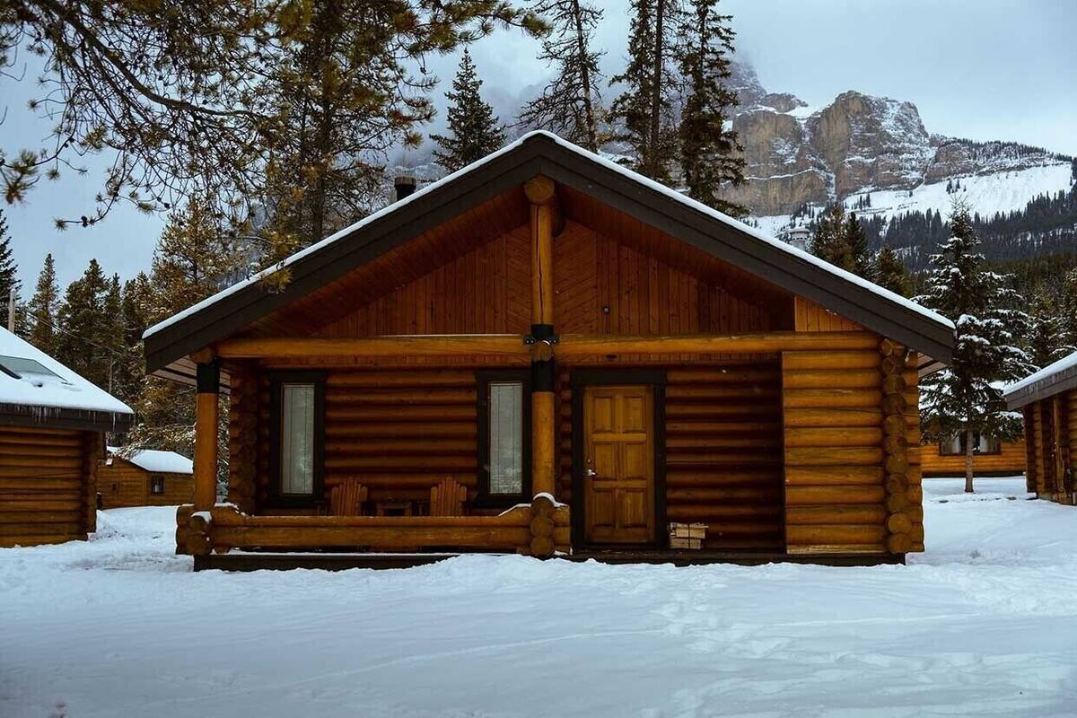 A log cabin with a pitched roof, large windows, and a door, surrounded by snow-covered ground and trees.