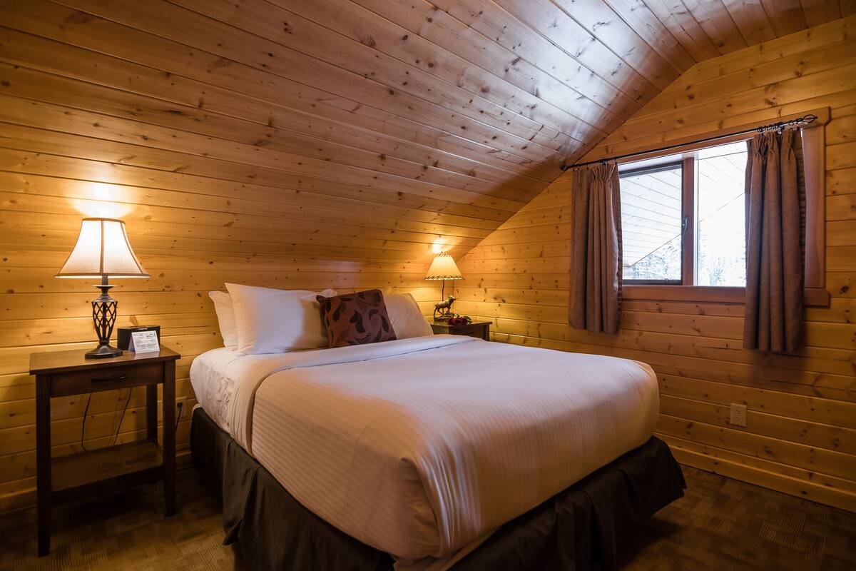 A cozy bedroom with a wooden ceiling, a large bed with white linens, two bedside tables with lamps, and a window with curtains.