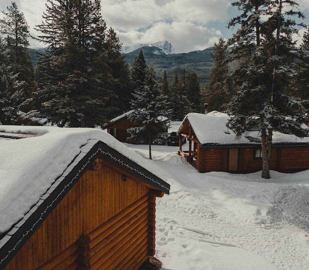 Een houten hut met een sneeuwbedekte dak, omgeven door hoge dennenbomen en een bergachtige achtergrond.