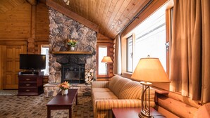 A log cabin interior with a stone fireplace, wooden furniture, a television, and a large window with curtains. - Castle Mountain Chalets (Castle Junction)
