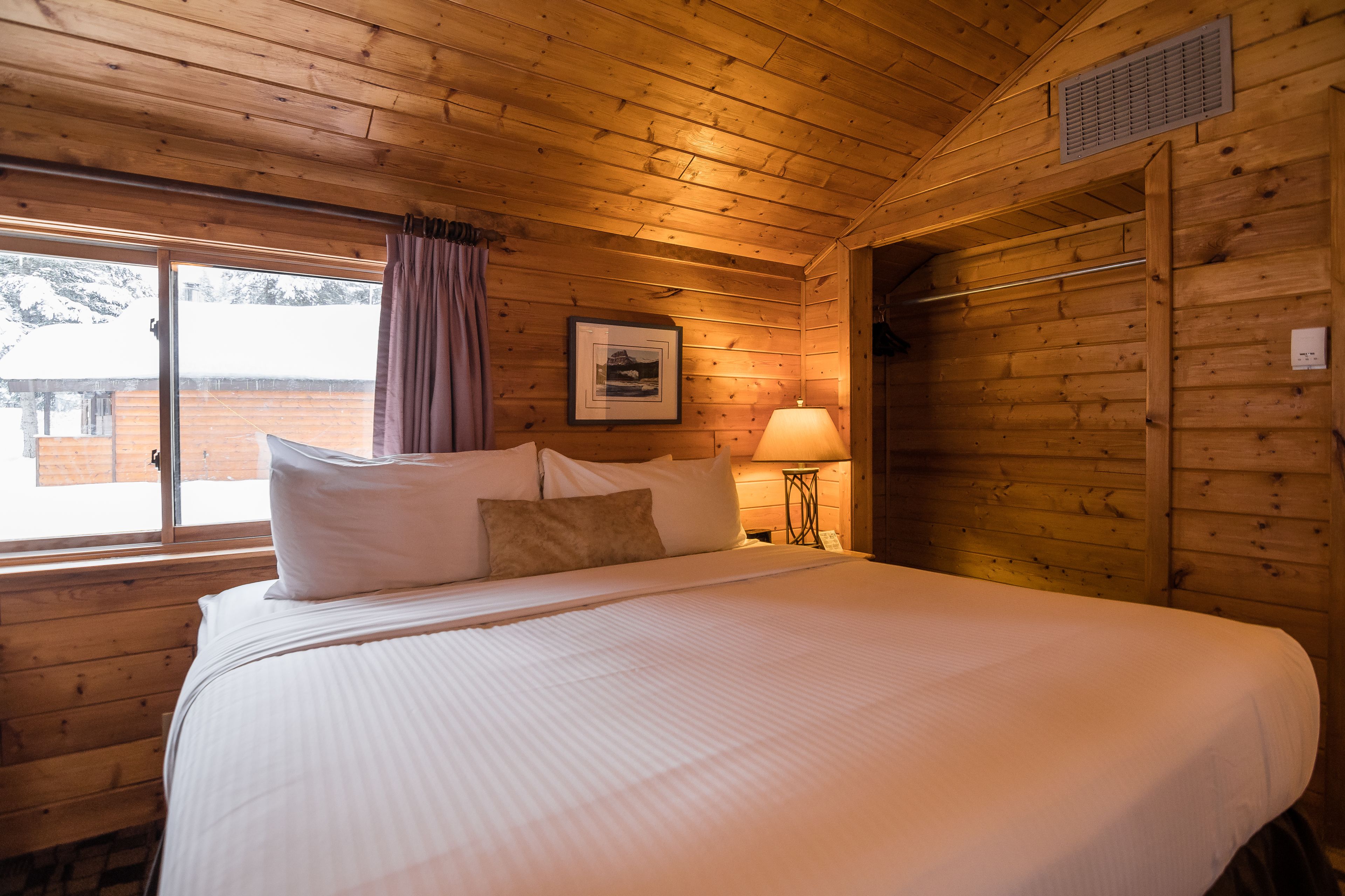 A wooden cabin room with a large bed, a window with curtains, a framed picture on the wall, and a lamp on a side table.