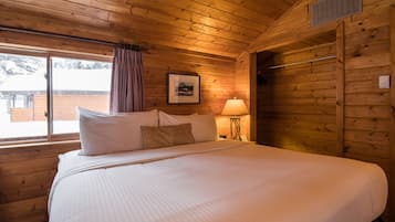 A wooden cabin room with a large bed, a window with curtains, a framed picture on the wall, and a lamp on a side table.