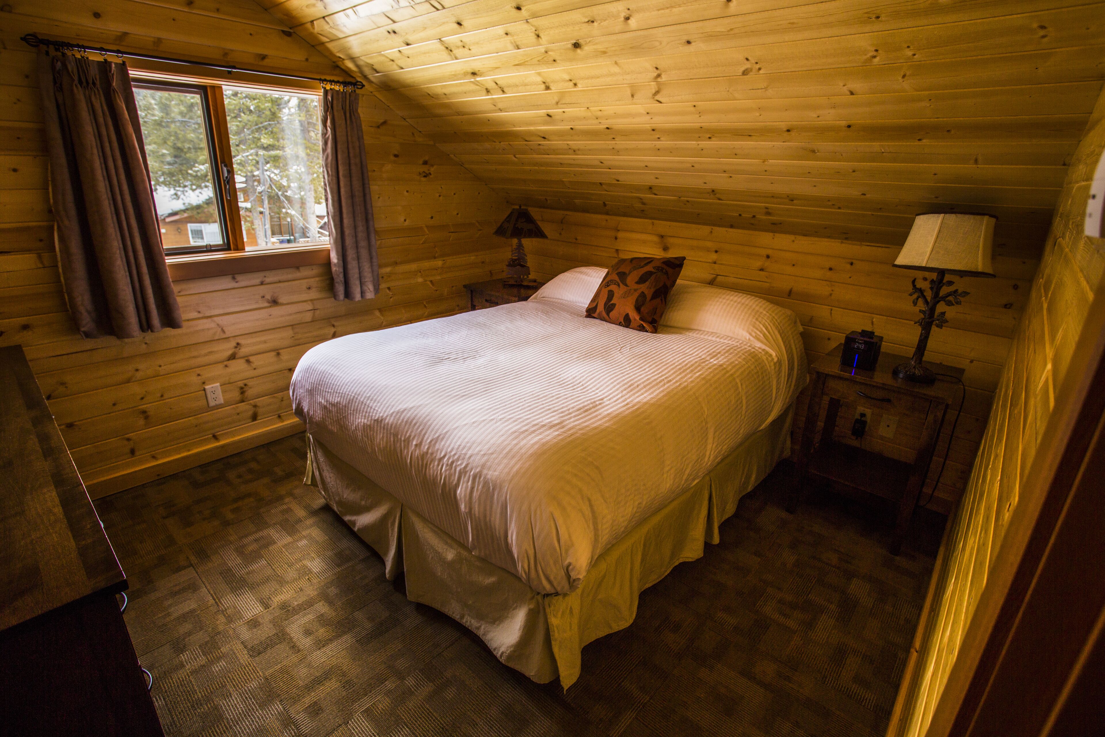 Un dormitorio rústico de cabaña con una cama, una mesita de noche, una lámpara y una ventana con cortinas.