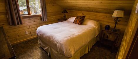Un dormitorio rústico de cabaña con una cama, una mesita de noche, una lámpara y una ventana con cortinas.