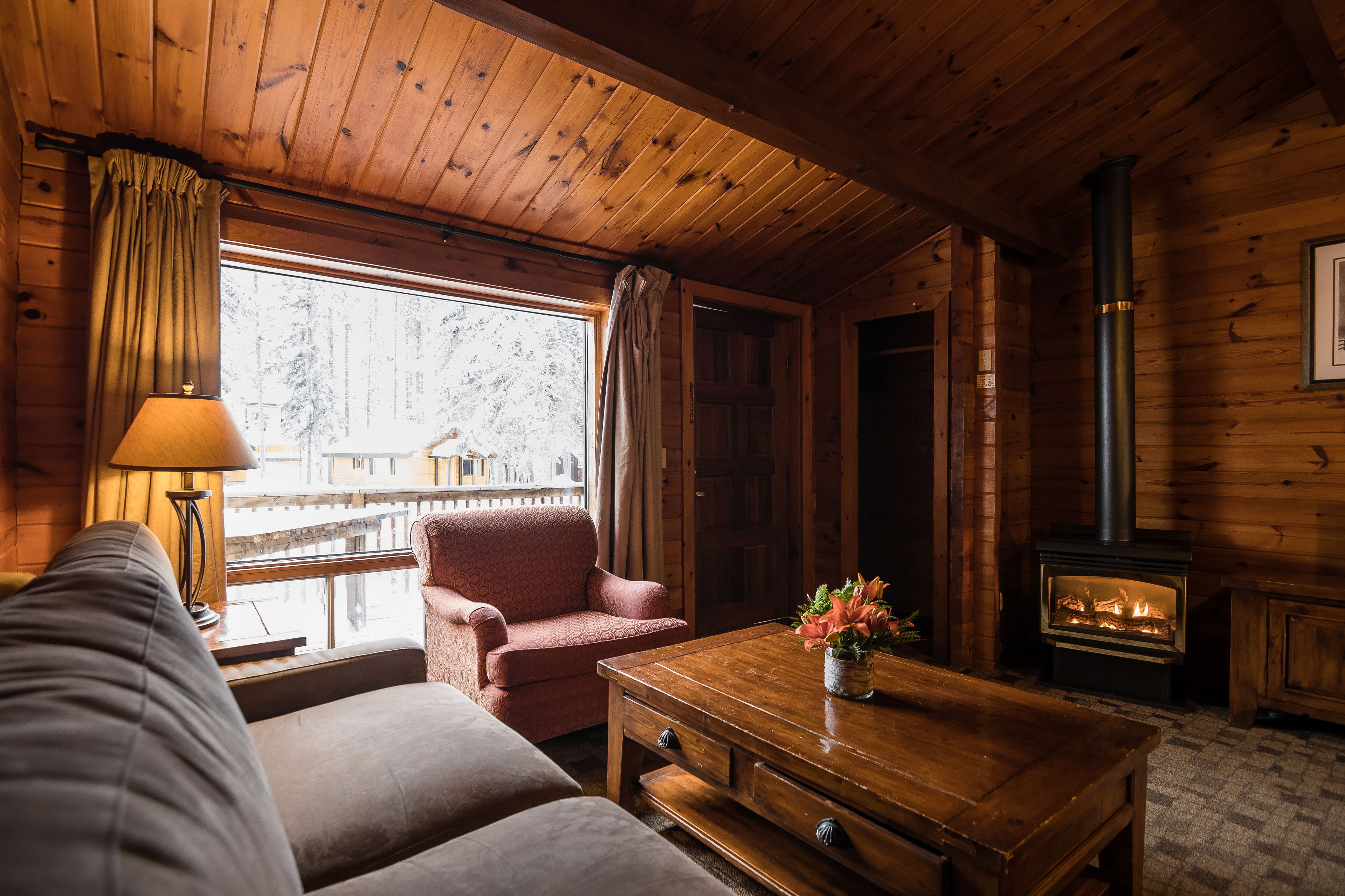 Un acogedor interior de cabaña con un sofá, una mesa de madera, una chimenea y una ventana con cortinas.