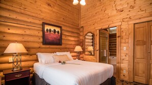 A log cabin bedroom with a large bed, two bedside lamps, a nightstand, a wooden door, and a framed picture on the wall. - Castle Mountain Chalets (Castle Junction)