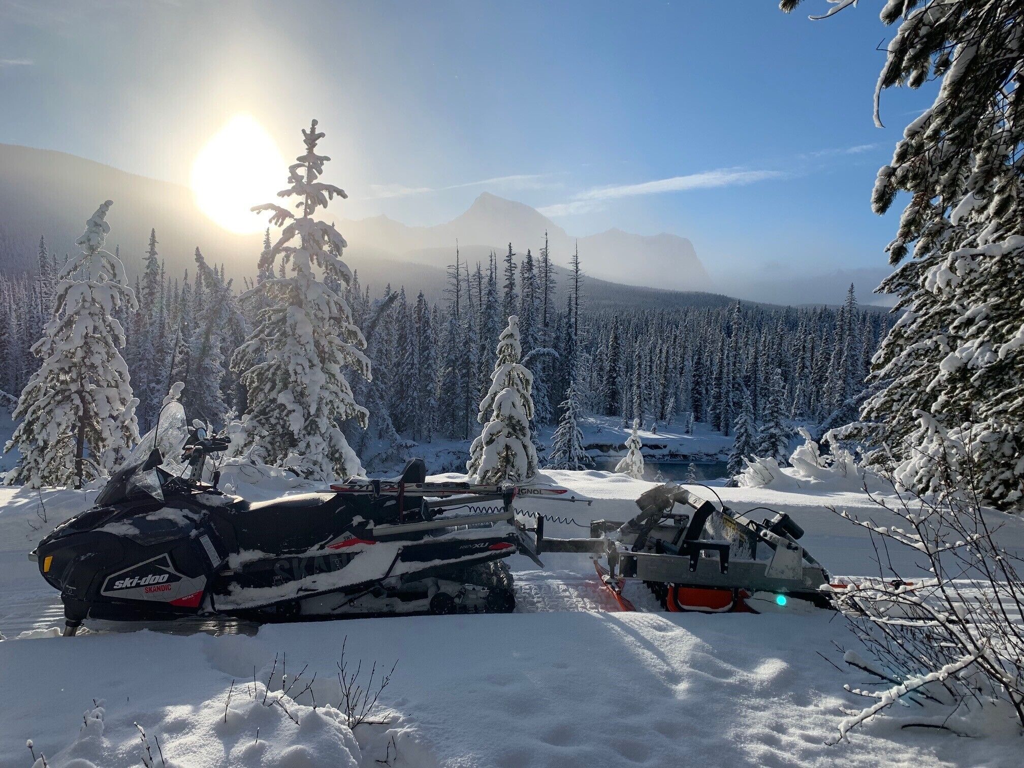 A snowmobile and a snowmobile trailer in a snowy forest with mountains in the background.