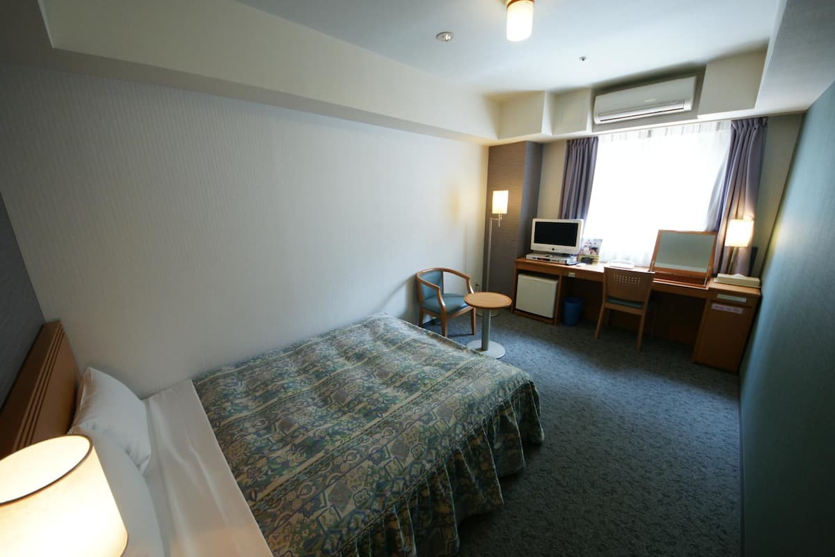 A hotel room with a single bed, a desk with a computer, a chair, and a small table.