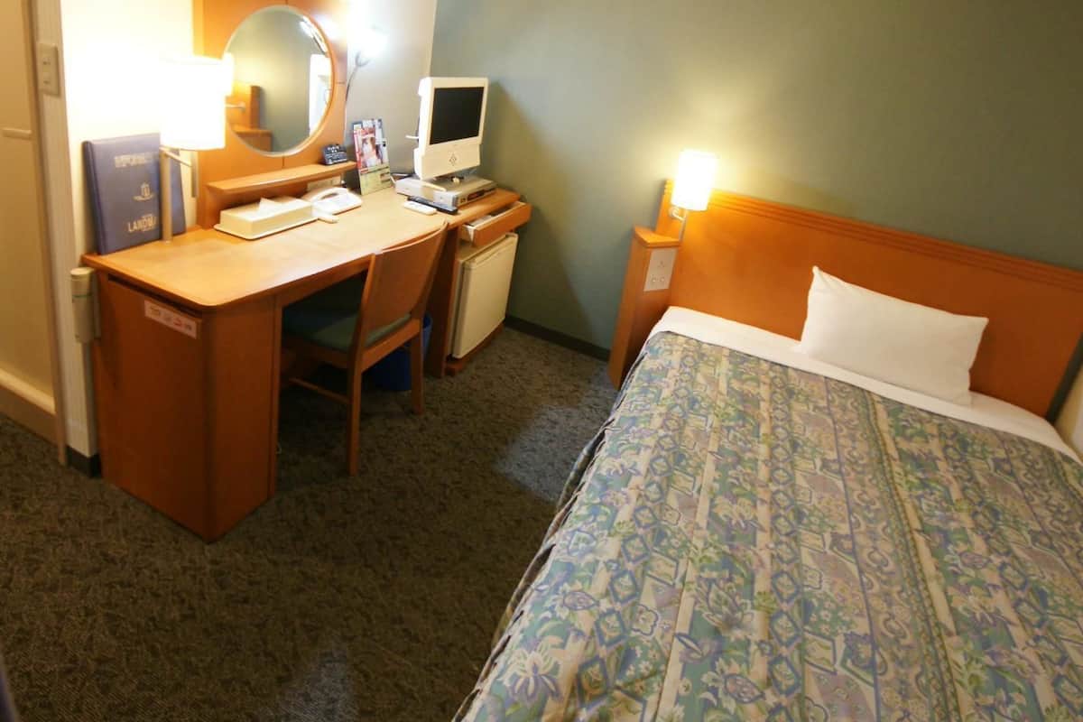 A hotel room with a bed, desk, chair, computer, and a lamp.