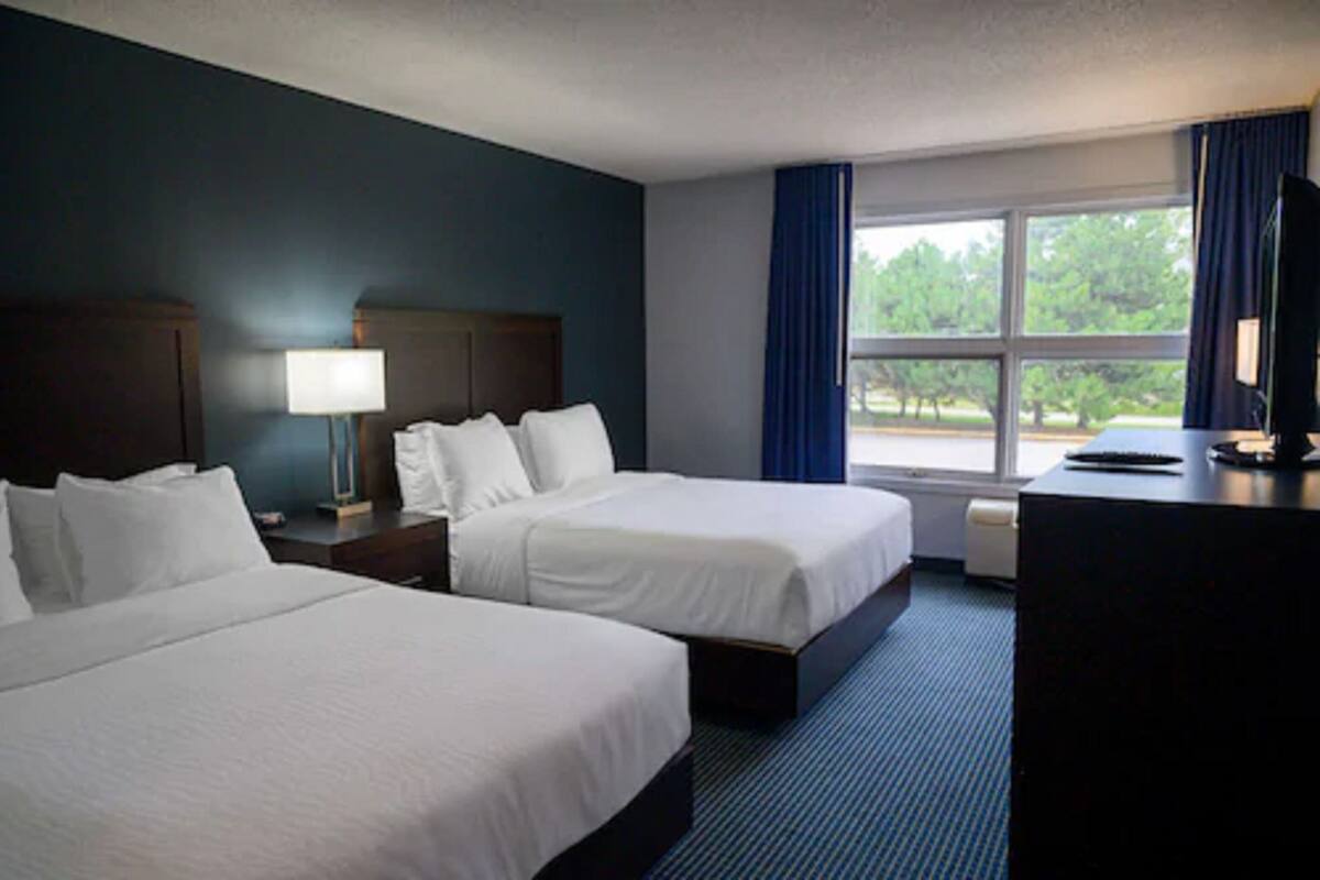A hotel room with two beds, a desk, a lamp, and a large window with curtains.