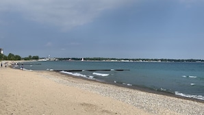 Une plage bordée d’une côte rocheuse, un phare visible au loin, et quelques personnes qui se promènent.