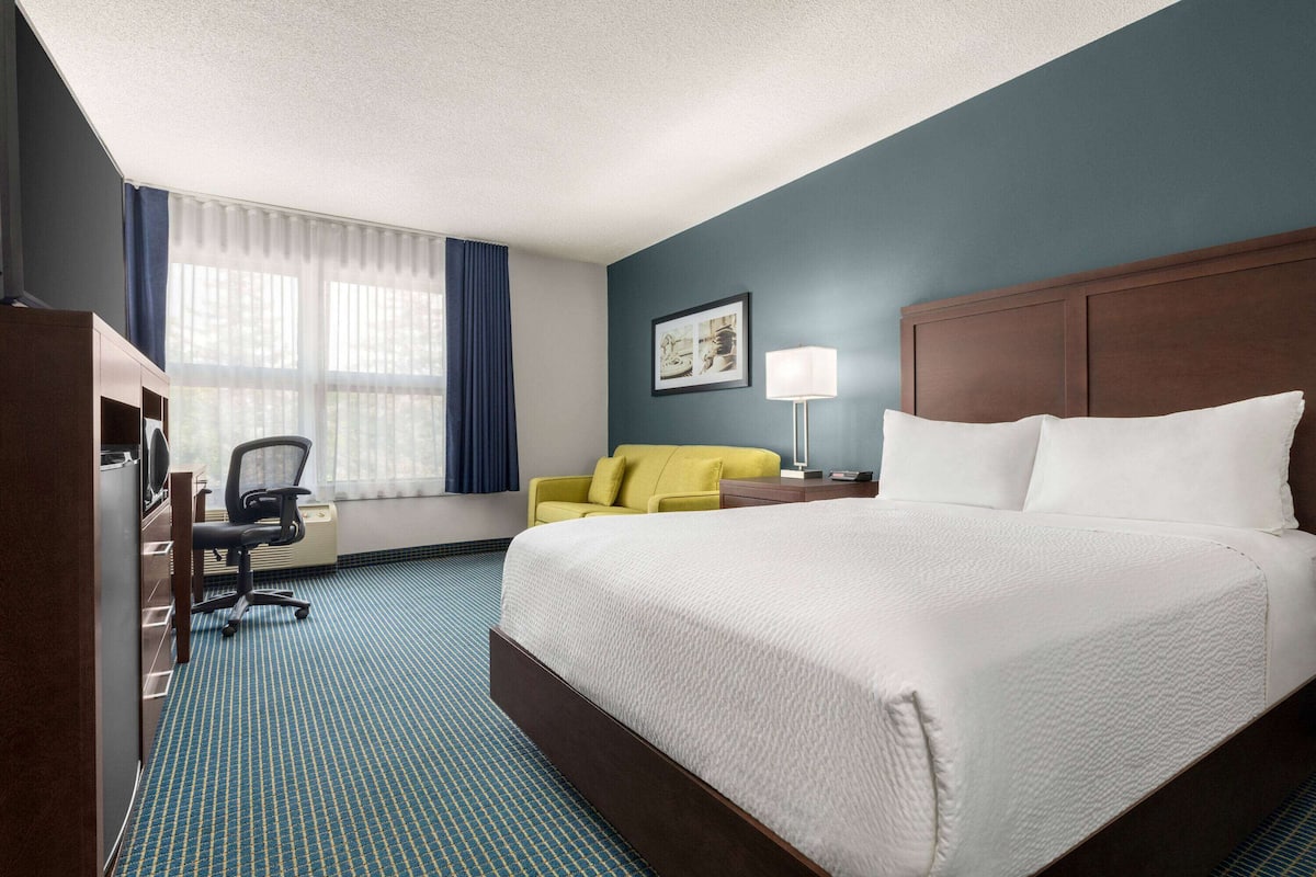 A hotel room with a large bed, a desk with a chair, a yellow armchair, a lamp, and a window with curtains.