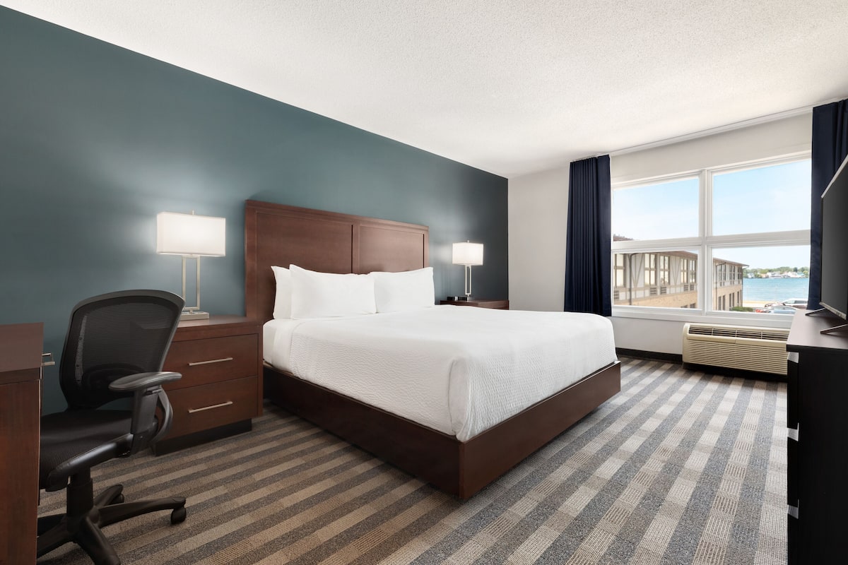 A hotel room with a large bed, two bedside tables with lamps, a desk with a chair, a view of the sea through a window, and a carpeted floor.