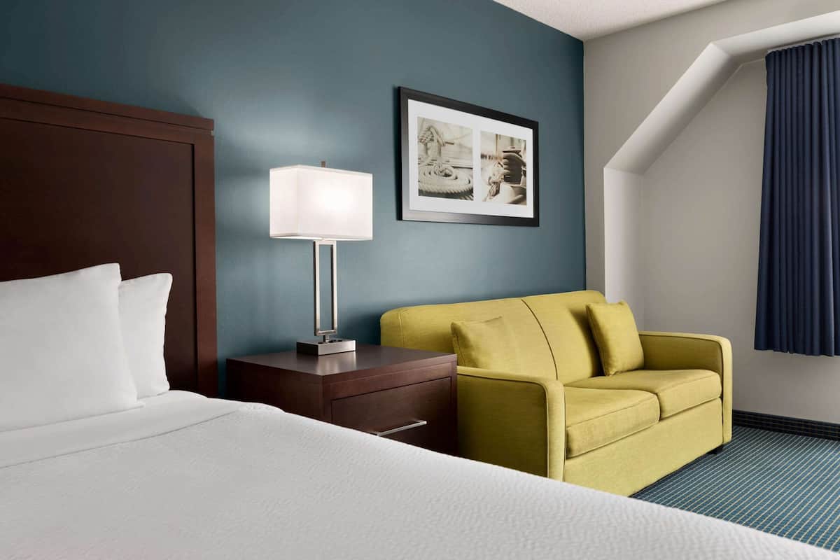A hotel room with a bed, a nightstand with a lamp, a yellow armchair, and a framed picture on the wall.