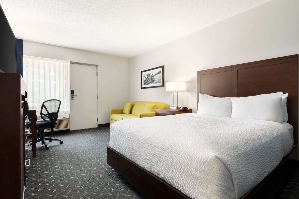 A hotel room with a large bed, a desk with a chair, a yellow sofa, and a window with blinds.