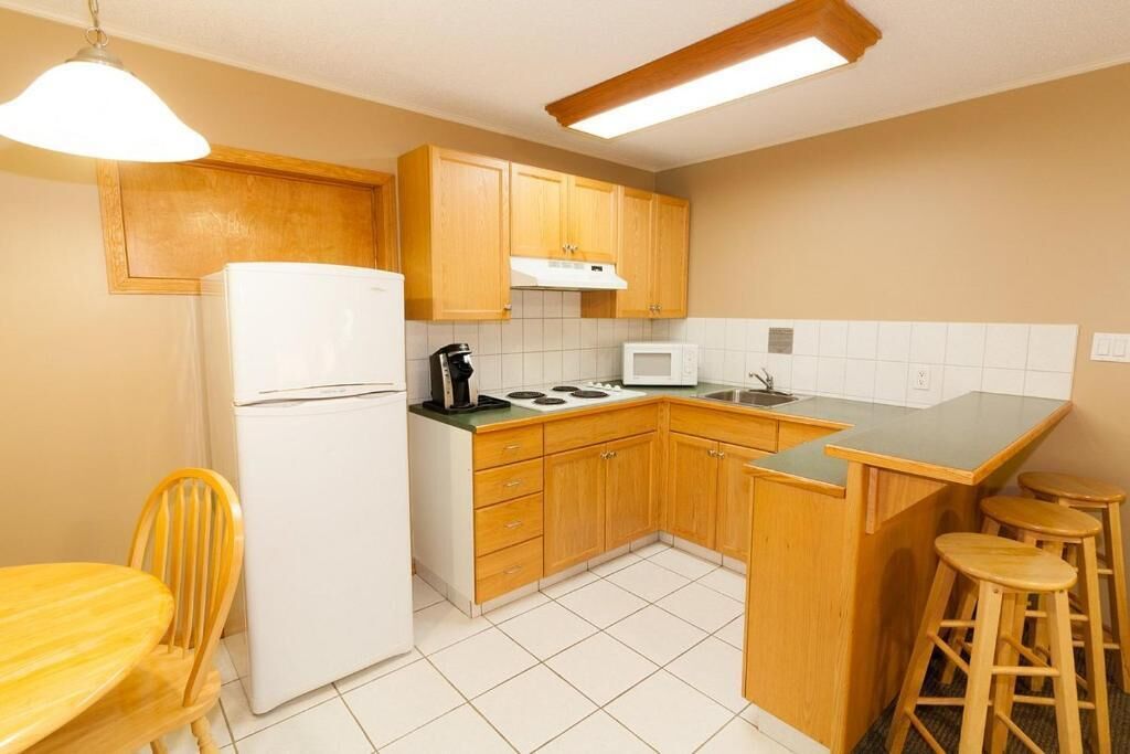A compact kitchen with wooden cabinets, a white refrigerator, a microwave, and a stove. There are two stools and a wooden table.