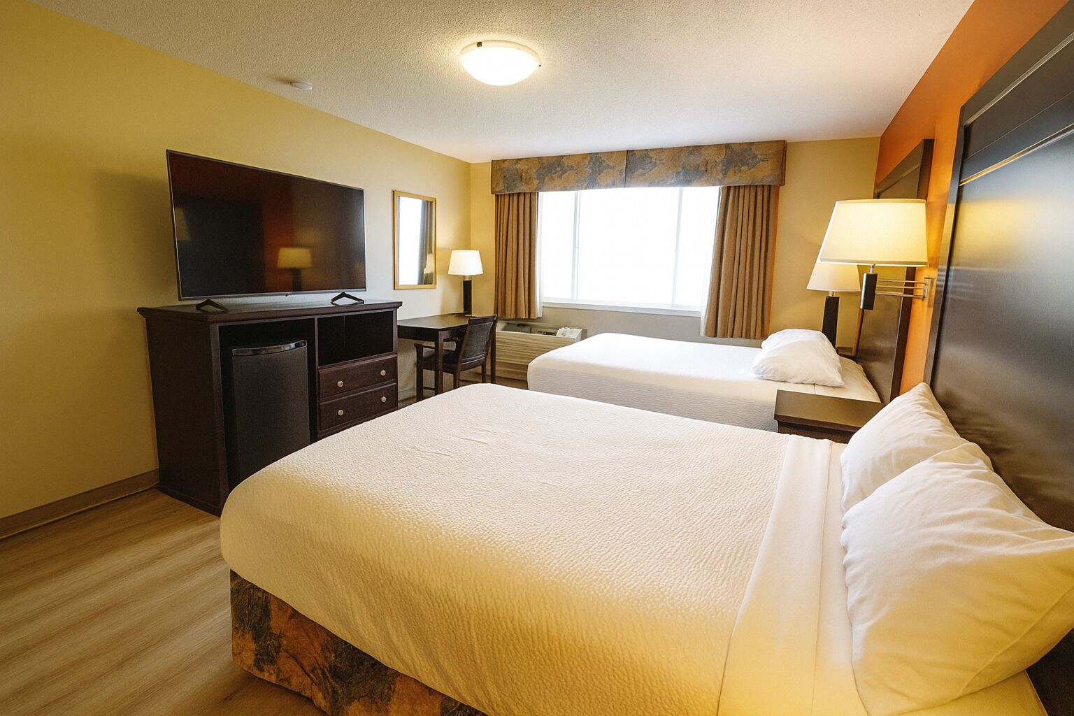 A hotel room with two beds, a TV, a desk, and a window with curtains.
