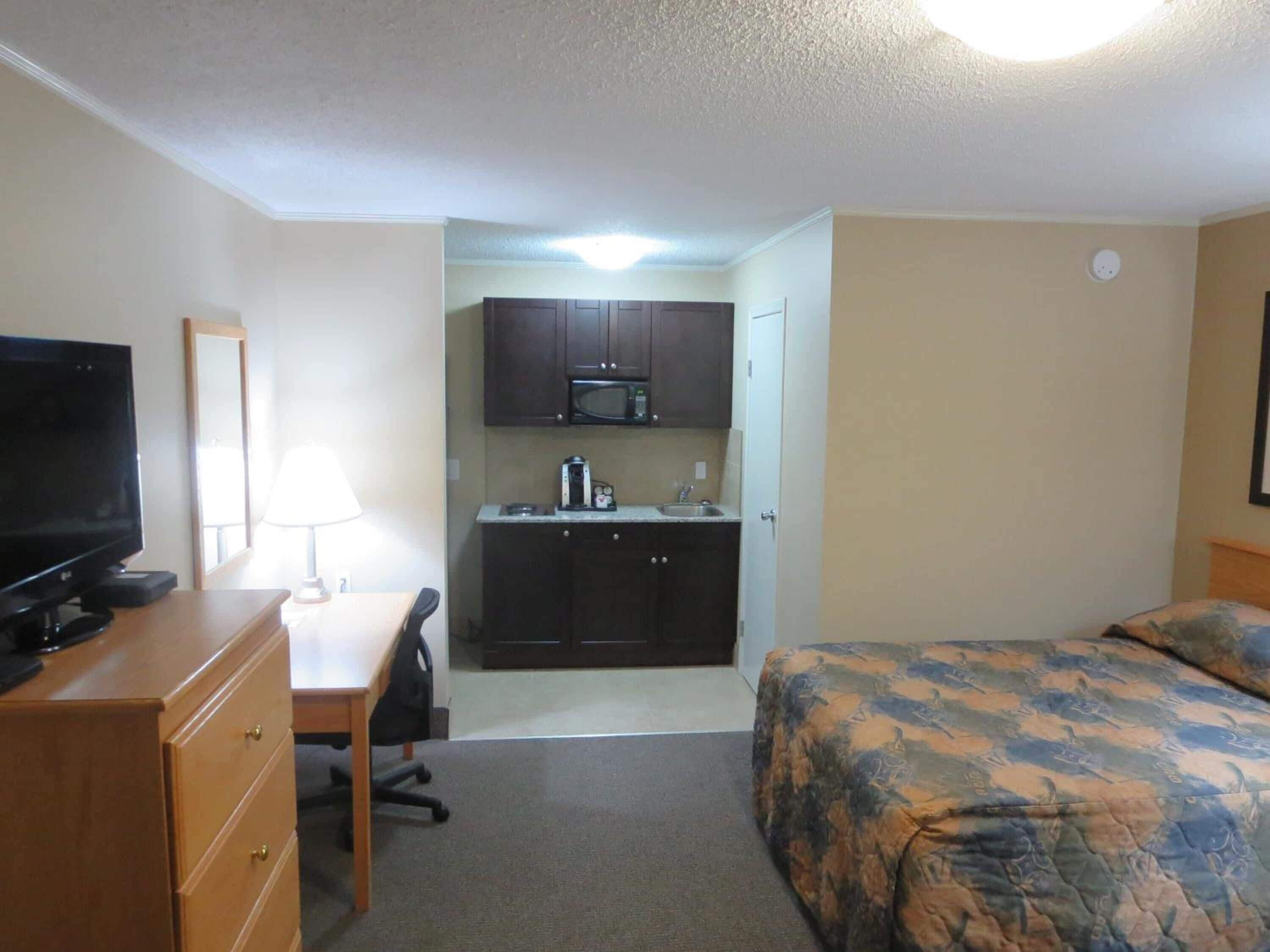 A compact hotel room with a bed, a desk with a TV, a kitchenette with a microwave, and a small office chair.