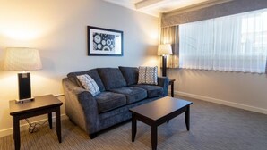A modern living room with a dark grey sofa, two side tables, a framed picture on the wall, and a large window with sheer curtains. - BCMInns Fort McMurray (Fort McMurray)