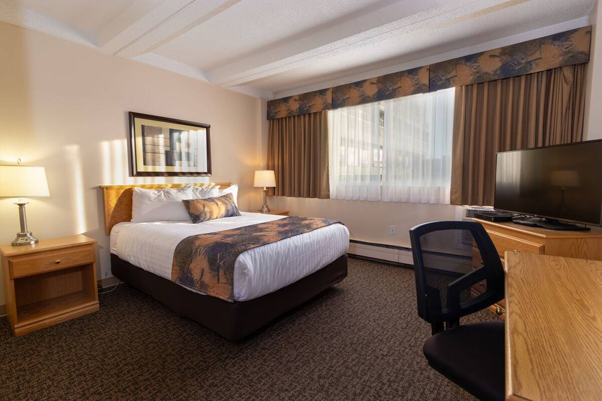 Habitación de hotel con cama, escritorio con silla, televisor y ventana con cortinas.