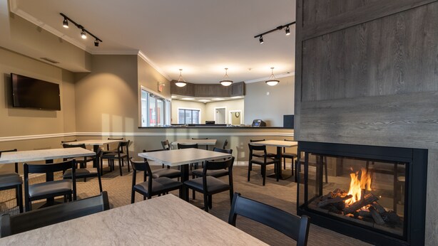 A modern dining area with tables and chairs, a fireplace, and a kitchen in the background. - BCMInns Fort McMurray (Fort McMurray)