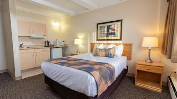 A hotel room with a bed, a nightstand, a lamp, a painting on the wall, and a kitchenette in the background.