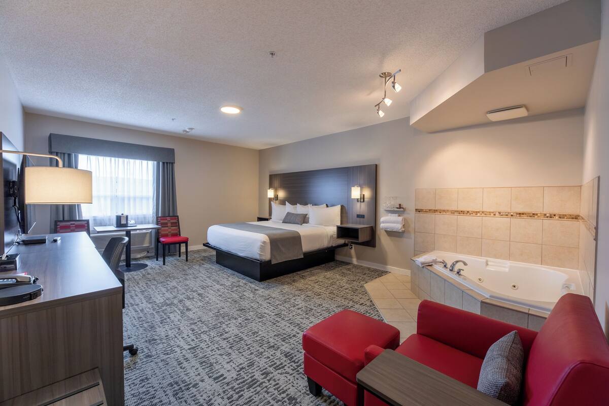 A hotel room with a bed, a desk, a red sofa, a bathroom with a bathtub, and a large window with curtains.