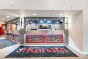 A reception area with a modern design, featuring a reception desk, a logo on the wall, and a carpet with the hotel name. - The Kanata by BCMinns Hinton (Hinton)