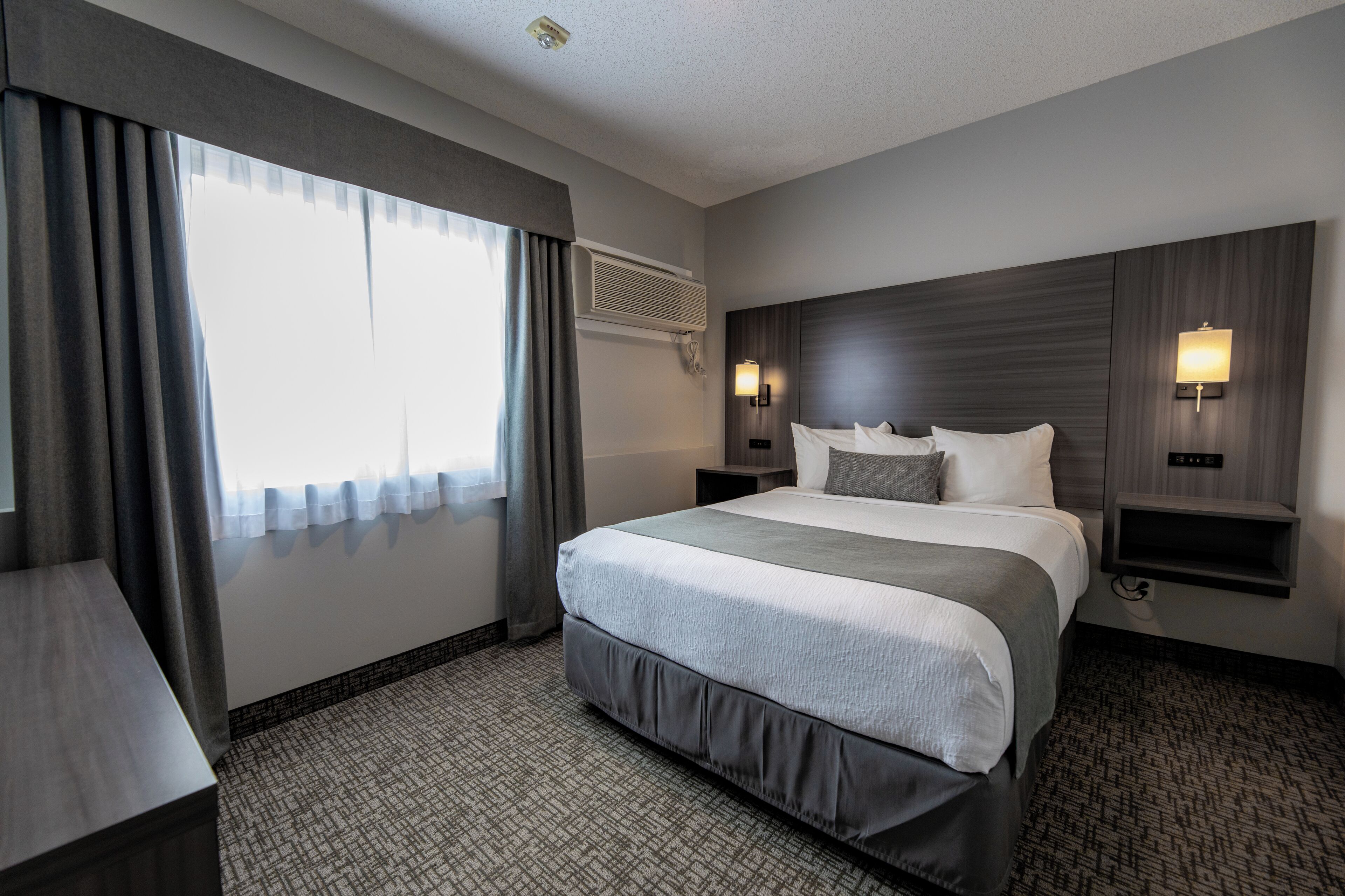 A hotel room with a large bed, a bedside table, a nightstand, a wall-mounted air conditioner, and a window with curtains.