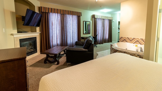 A hotel room with a large bed, a desk, a chair, a fireplace, a television, and a bathtub. - BCMInns Lac La Biche (Lac La Biche)