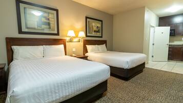 A hotel room with two beds, a wooden headboard, wall-mounted lamps, and a kitchen area visible through an open door.