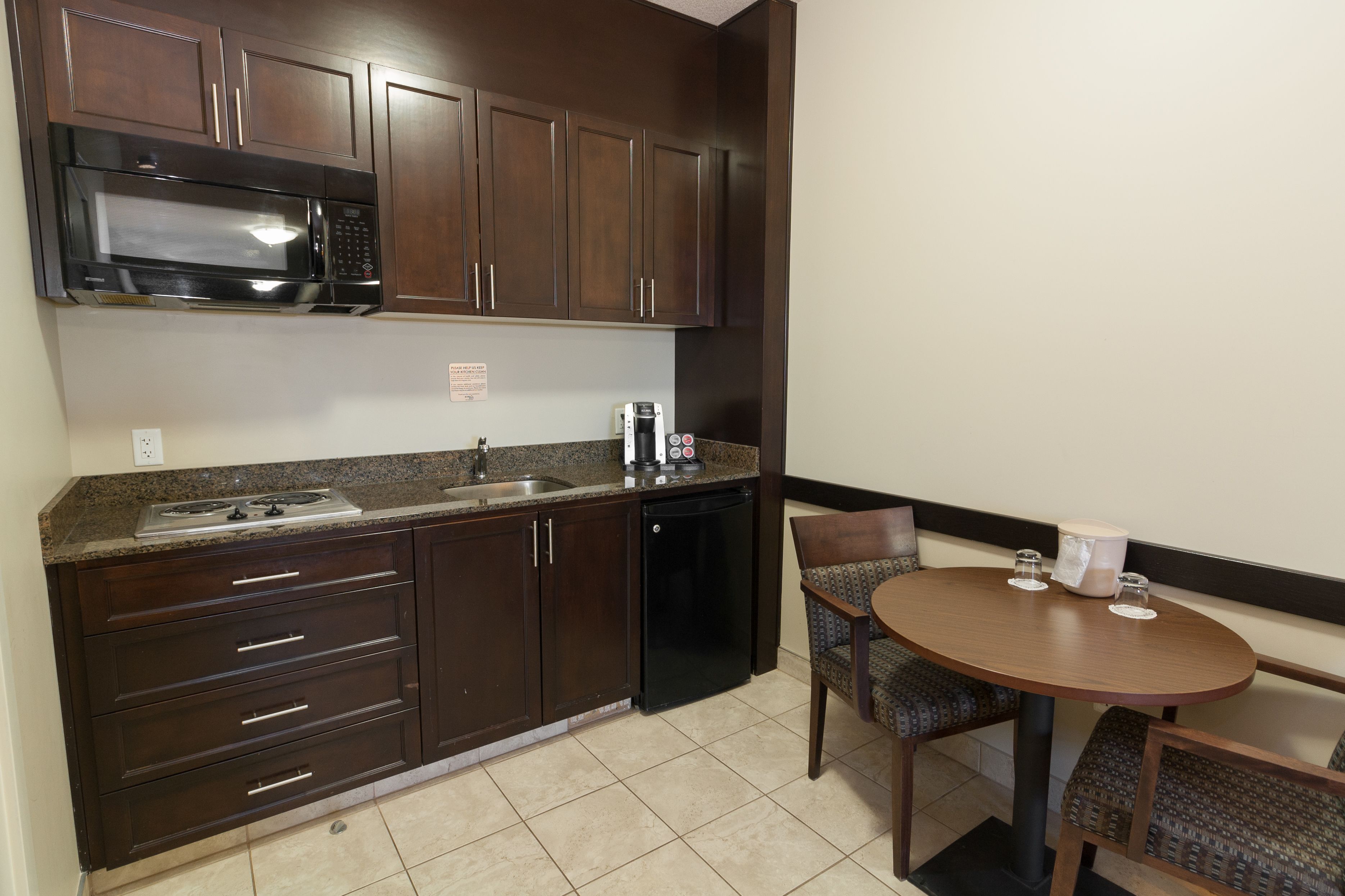 A compact kitchen with dark wooden cabinets, a stainless steel microwave, a built-in dishwasher, a granite countertop, and a small round table with two chairs.