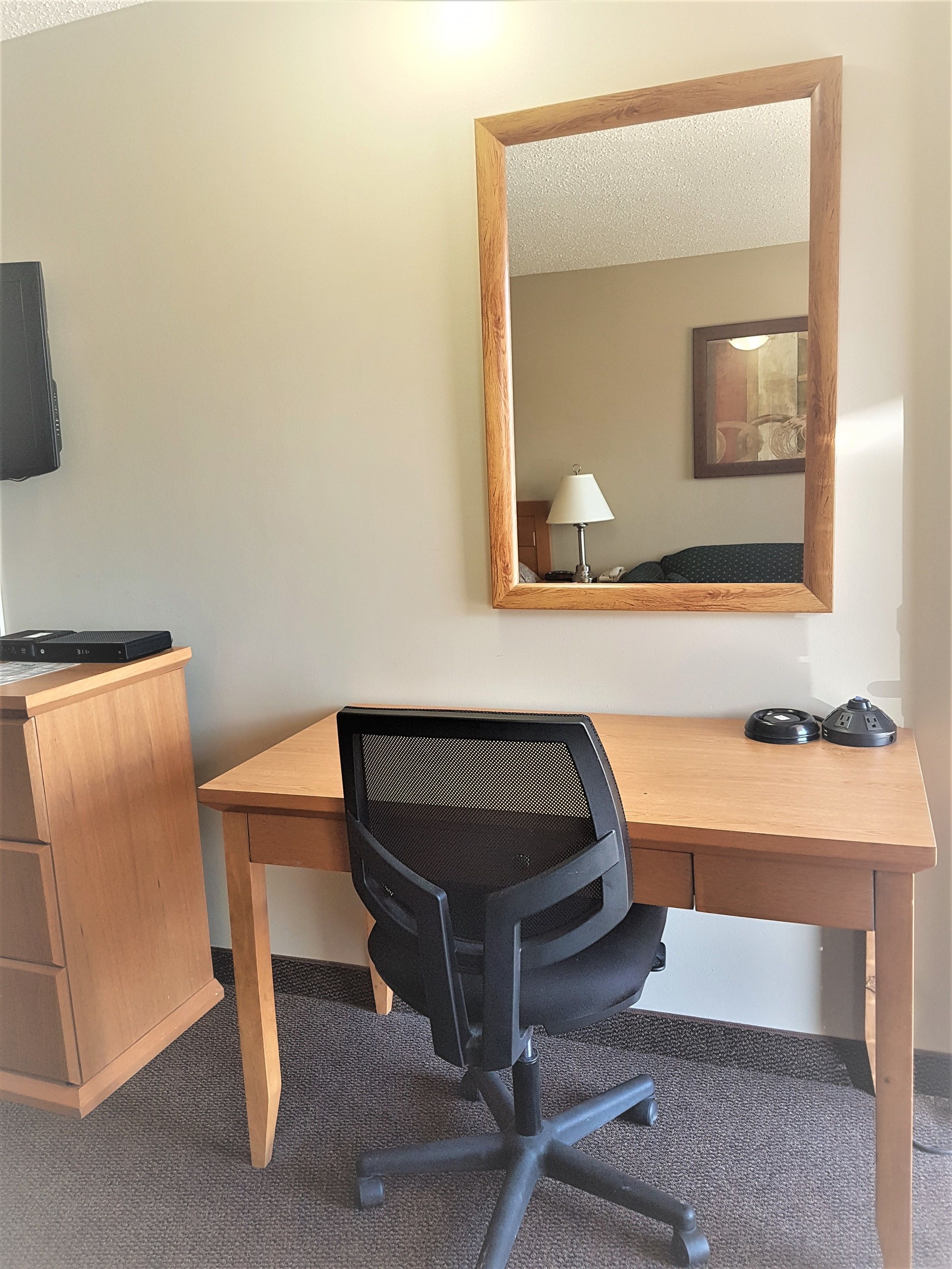 A hotel room with a wooden desk, a black office chair, a mirror, a lamp, and a television.