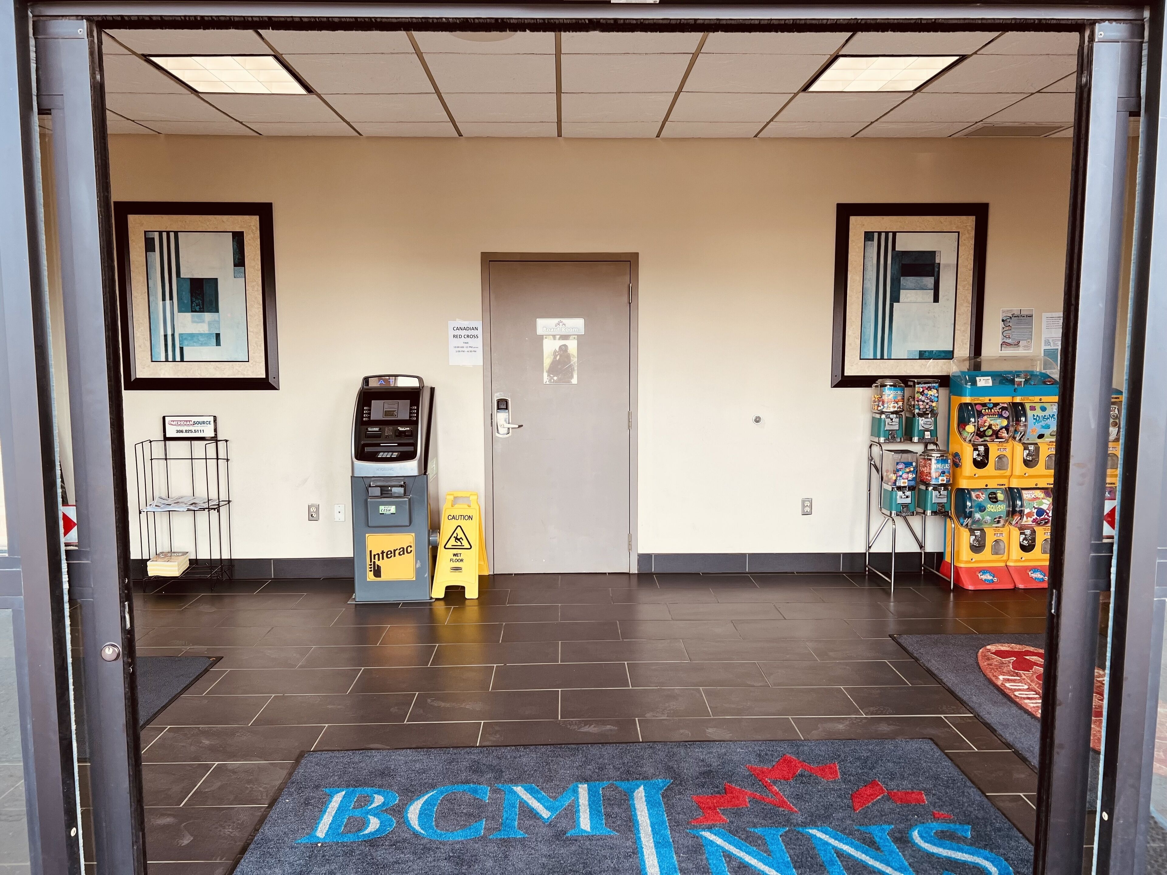 A hotel entrance with a wet floor sign, a self-service kiosk, vending machines, and a doormat with "BCMT INNS" text.