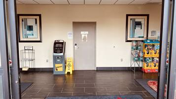 A hotel entrance with a wet floor sign, a self-service kiosk, vending machines, and a doormat with "BCMT INNS" text.