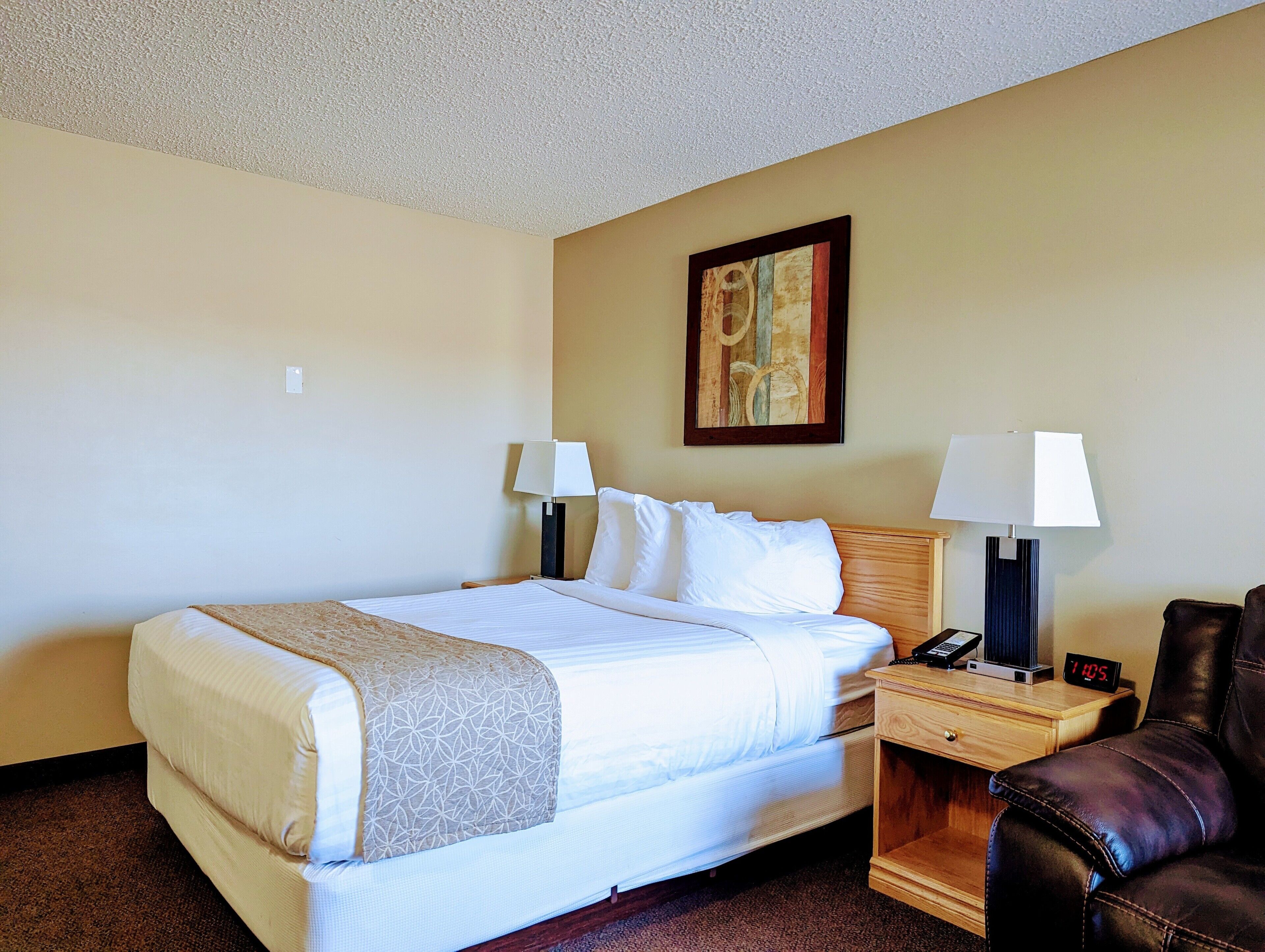 A hotel room with a bed, two bedside lamps, a clock, a leather chair, and a painting on the wall.