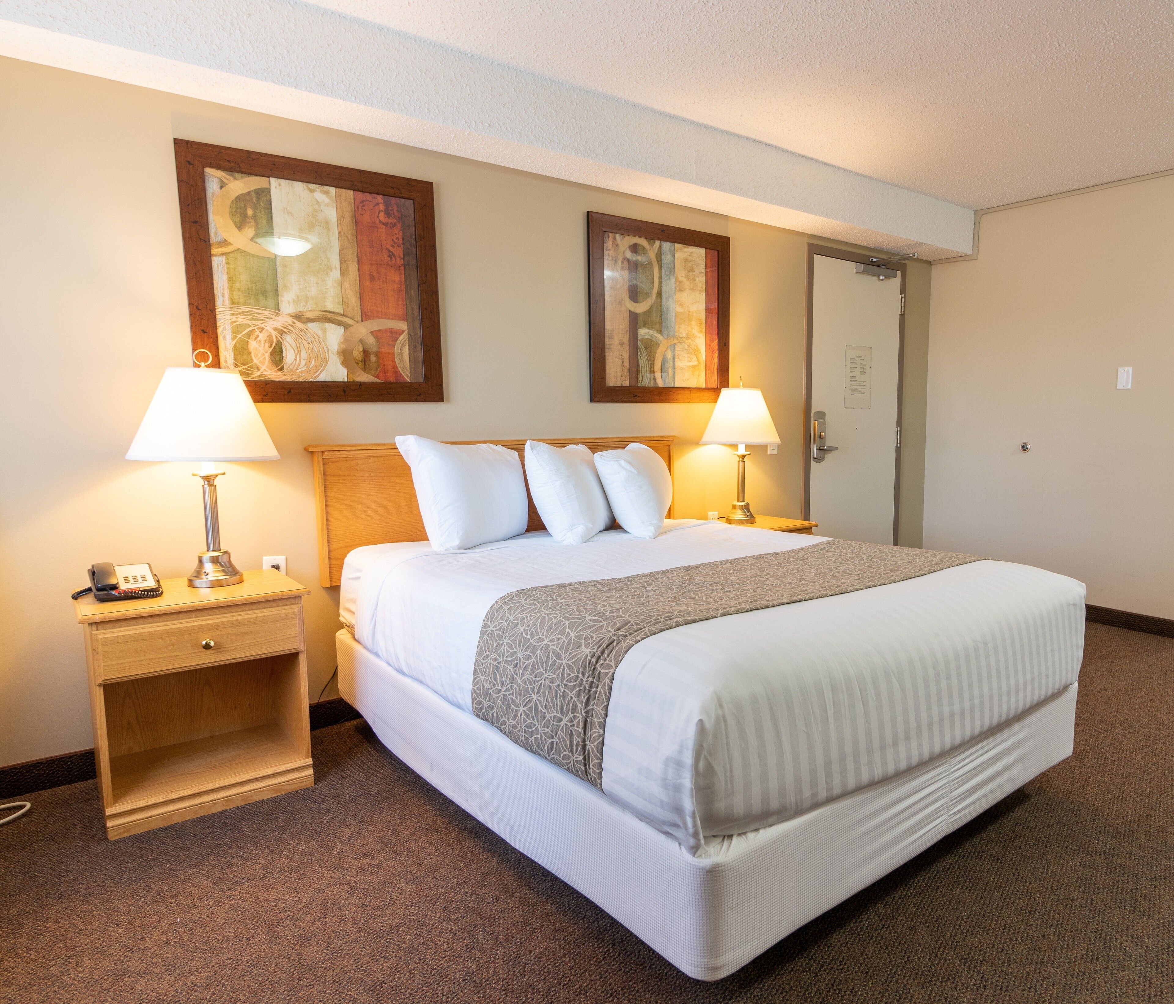 A hotel room with a bed, two bedside lamps, a nightstand, a door, and two framed artworks on the wall.