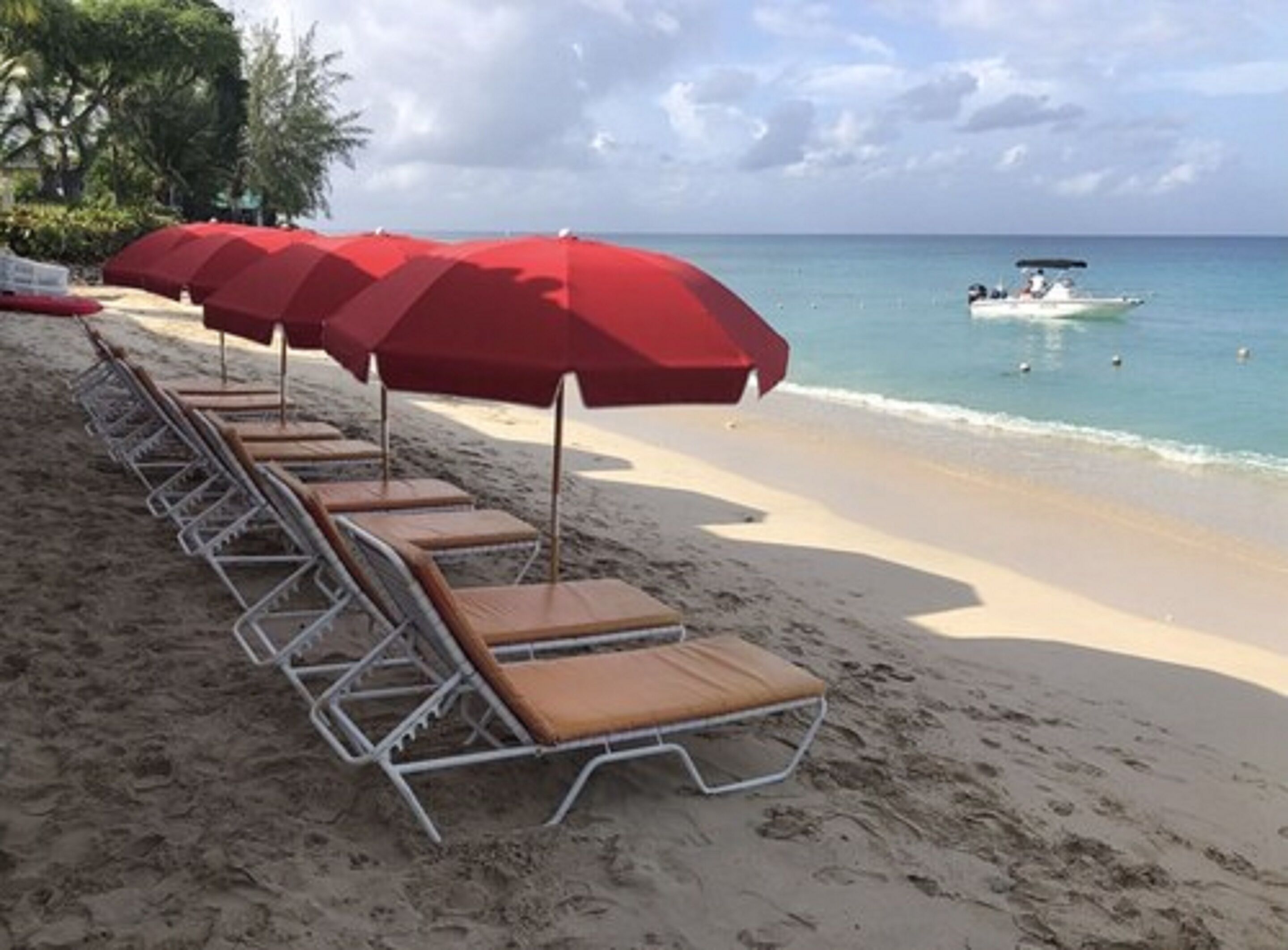Una playa con filas de tumbonas y sombrillas rojas, un barco en el agua, y un cielo despejado.
