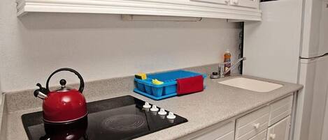Una cocina con una placa de inducción negra, un hervidor rojo, un microondas y un refrigerador blanco.