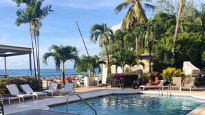 A pool area with lounge chairs, palm trees, and a view of the ocean. - Tropical Sunset Beach Apartment Hotel (Holetown)