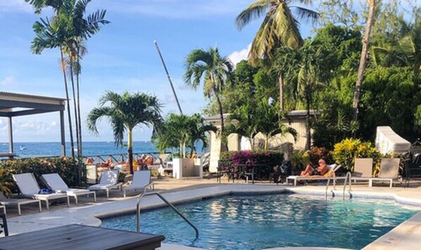 A pool area with lounge chairs, palm trees, and a view of the ocean. - Tropical Sunset Beach Apartment Hotel (Holetown)