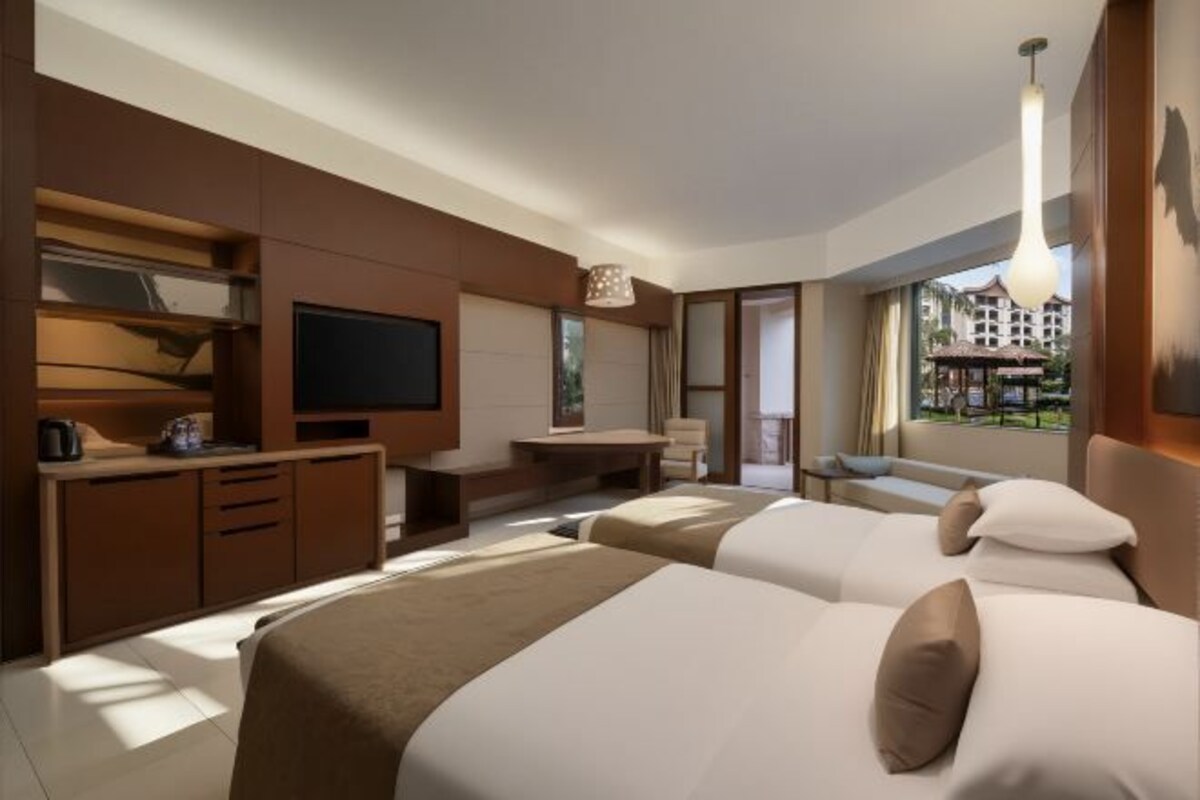 A hotel room with two beds, a TV, a desk, and a view of a building.