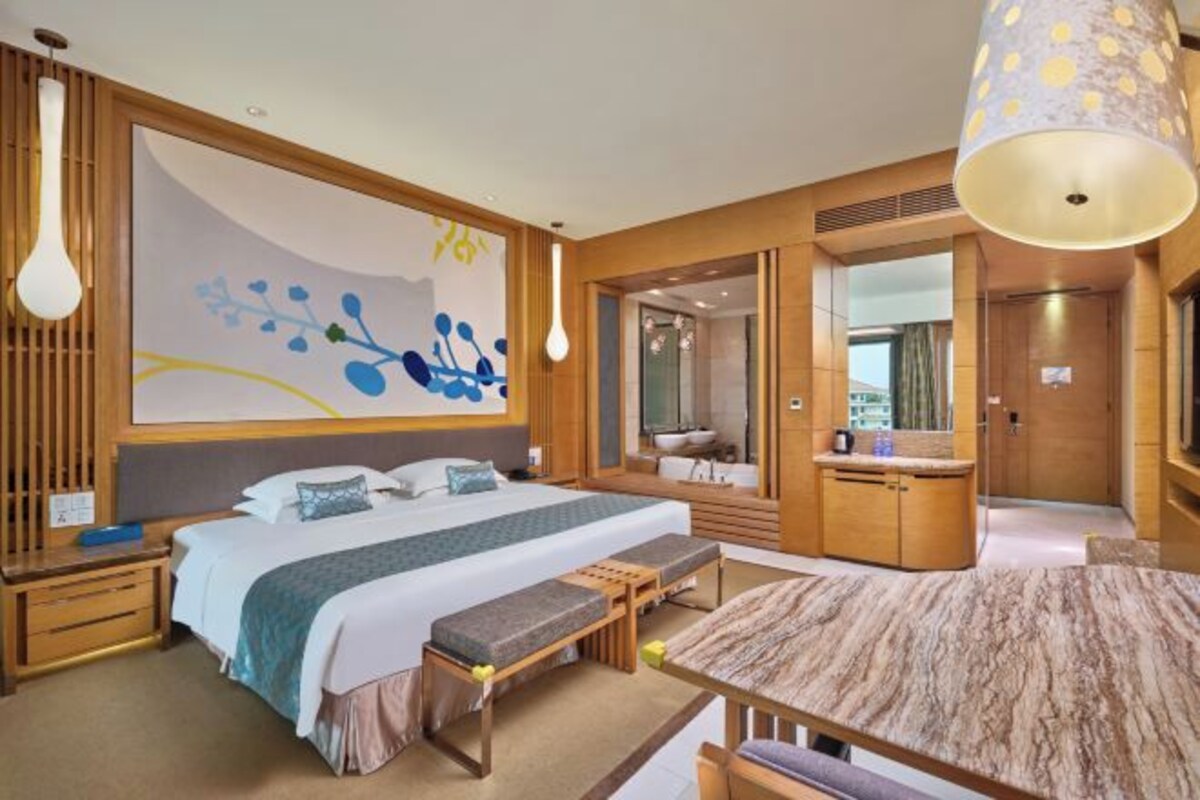 A modern hotel room with a large bed, a wooden bench, a dining table, and a large window.